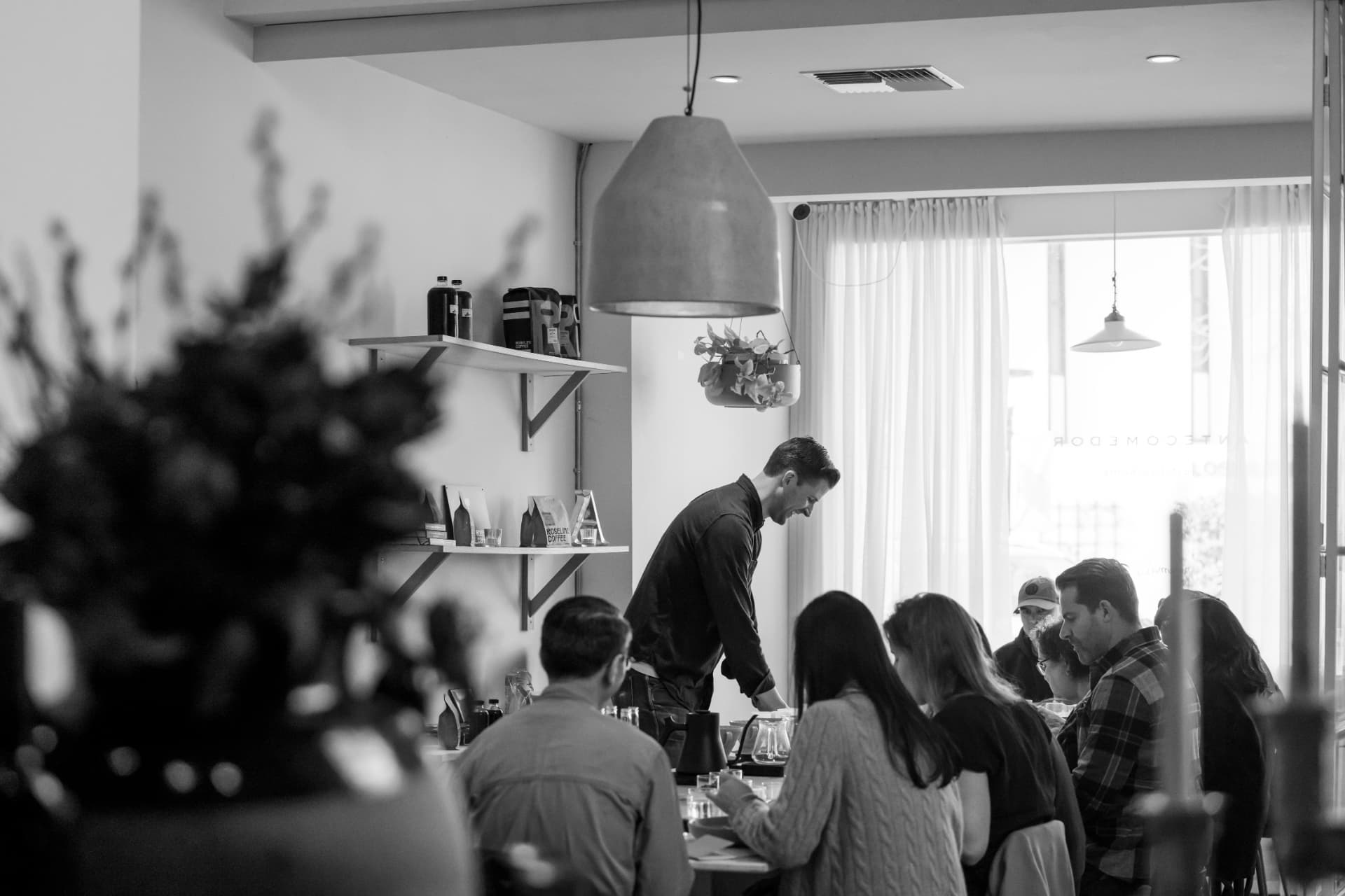 A black and white photograph of a barista serving guests seated around a table in a sunlit tasting room